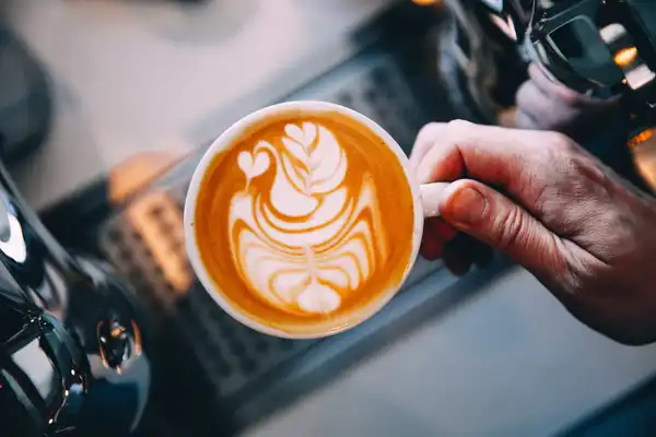 A hand holding a cappuccino with leaf-shaped latte art beside an espresso machine